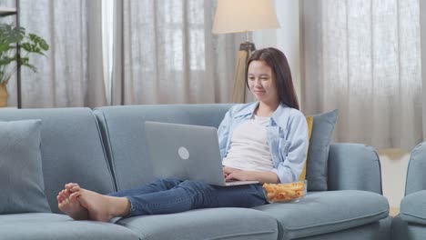 full body of asian teen girl with snacks typing on a laptop while lying on sofa in the living room at home