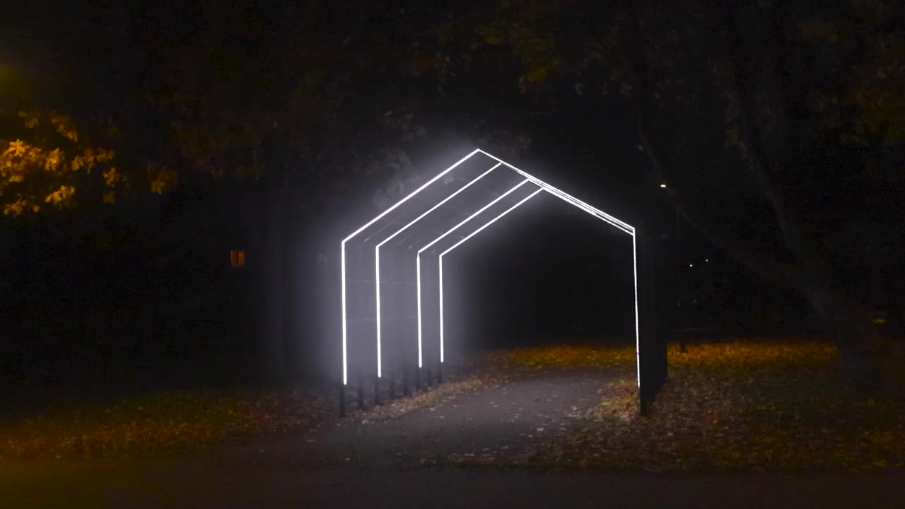 Artistieke en door design geleide verlichting die leidt naar een park dat gloeit tijdens een donkere herfstnacht. De lichten zijn helder en wit van kleur en gevormd als een huis of een driehoek, terwijl er gevallen bladeren op de grond liggen.