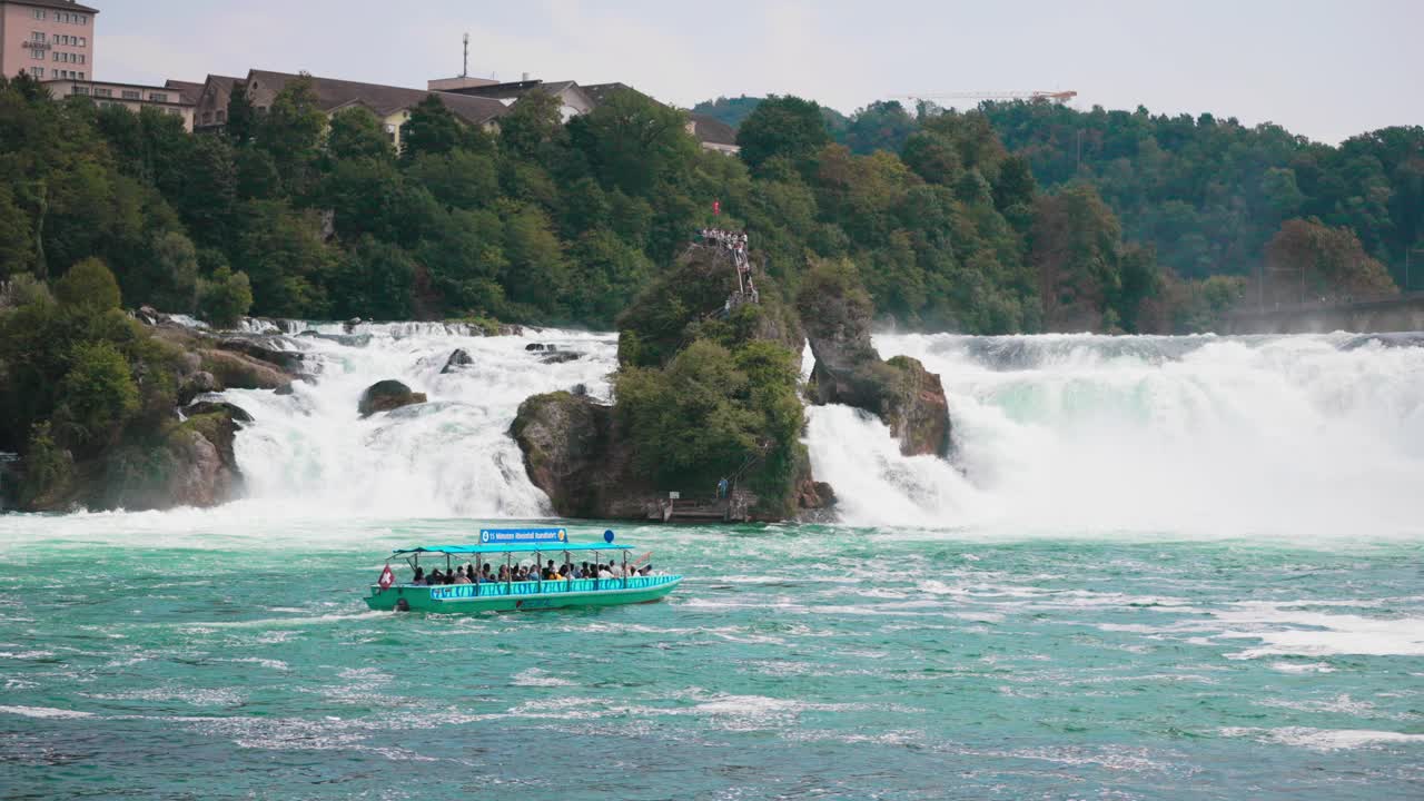 Thuyền du lịch chở đầy hành khách đi ngang qua thác Rhine ở Thụy Sĩ với đài quan sát bằng đá và rừng cây xanh tốt làm nền.