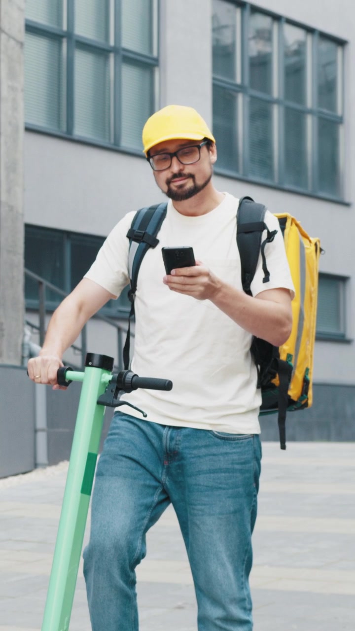 A delivery person wearing a yellow cap and backpack uses a smartphone while riding an electric