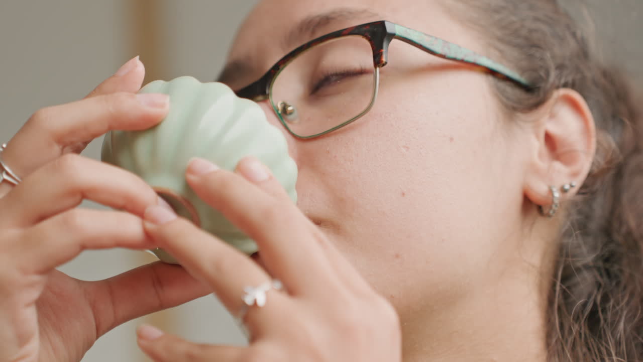 Primo piano in una camera d'hotel: una donna annusa la tazza, l'ospite assapora il delicato aroma del tè in una tazza di ceramica. Gli occhiali riflettono la luce della finestra, gioielli discreti all'orecchio e un anello, una quieta introspezione, una pausa lenta e consapevole.