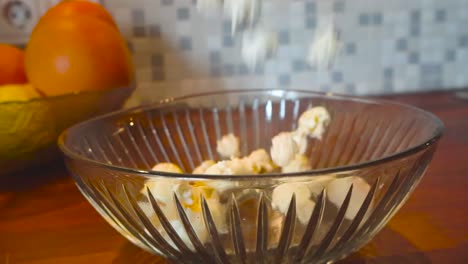 Close-up-slow-motion-footage-of-popcorn-being-poured-into-a-glass-bowl-on-a-kitchen-table-with-oranges-and-lemons-in-a-bowl-in-the-background