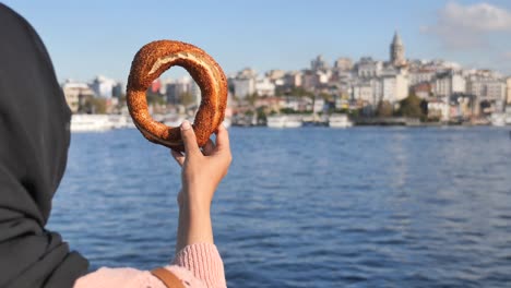mujer en hijab sosteniendo un simit con la torre de galata en el fondo