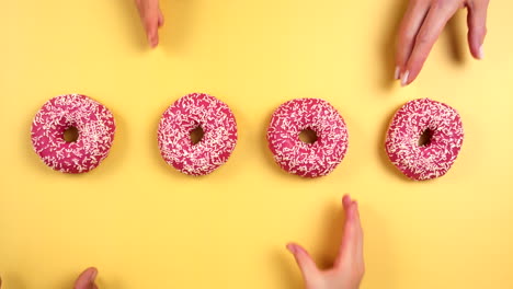 hands grabbing four pink colorful donuts on light yellow background. family take delicious donut at same time. child, male, female. pink glazed with sprinkles. filmed in prores, topview, slowmotion