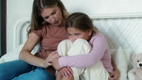 Mother-consoling-elementary-age-girl-sitting-on-bed-with-head-hidden-in-legs