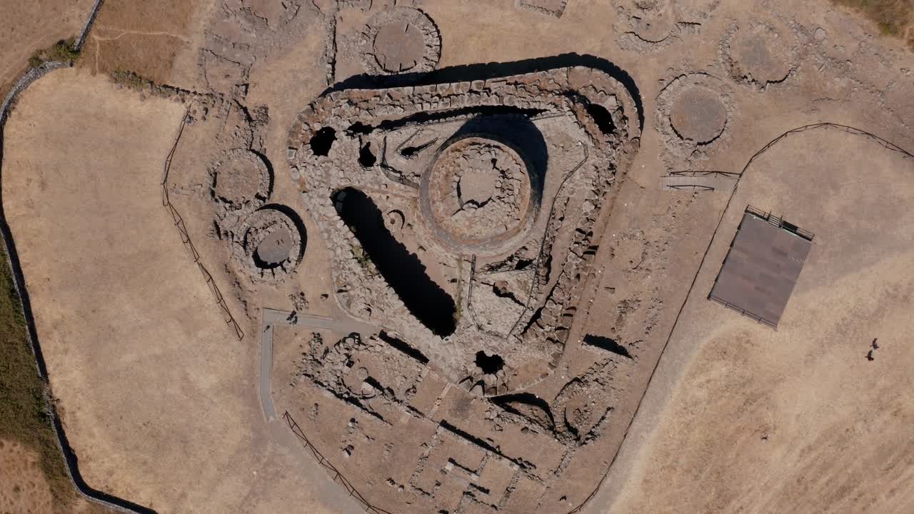 Top-down aerial drone shot of ancient Nuraghe stone complex showing triangular layout and circular structures surrounded by dry land in Barumini Sardinia Italy under bright daylight