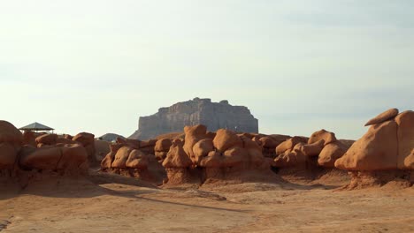 Magnífica-E-Impresionante-Toma-Inclinada-Hacia-Arriba-Del-Hermoso-Parque-Estatal-Goblin-Valley,-Utah,-Formaciones-Rocosas-De-Hongos-Con-Una-Gran-Montaña-De-Roca-Roja-En-El-Fondo-En-Un-Cálido-Y-Soleado-Día-De-Verano.