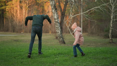 a man in a green jacket and a little girl in a pink jacket, both wearing blue jeans, are seen joyfully jumping and playing together outdoors on a lush green grassy field