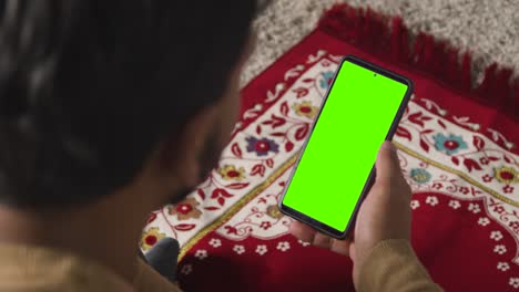 close up of muslim man at home sitting on floor on prayer mat holding green screen mobile phone