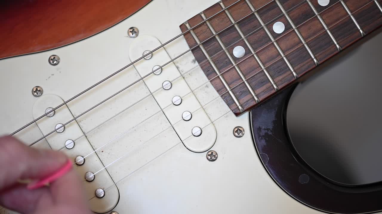 video de 4k de un primer plano de la mano de un hombre arrancando las cuerdas de una guitarra eléctrica con un plectrum rosa