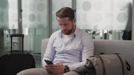 a young man consults his cell phone while waiting for his flight to depart in an airport lounge.