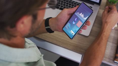 hombre caucásico sentado en un escritorio usando un teléfono inteligente con estadísticas en la pantalla