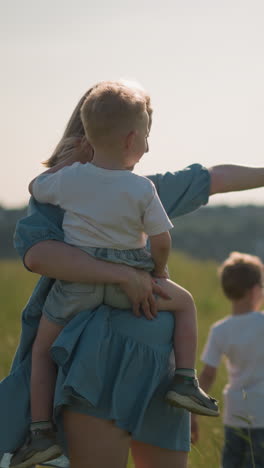 una mujer con un vestido azul lleva a su hijo pequeño mientras su hermano está cerca. los tres están apuntando hacia el lago, disfrutando de la vista panorámica. en un campo soleado y cubierto de hierba