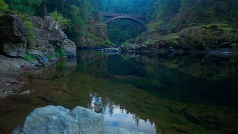 Eine-Bogenbrücke-überspannt-Einen-Sanft-Fließenden-Fluss-Im-Wald,-Verschmilzt-Mit-Dem-üppigen-Grün-Und-Schafft-Eine-Malerische,-Ruhige-Szenerie-Natürlicher-Schönheit-Und-Heiterer-Umgebung