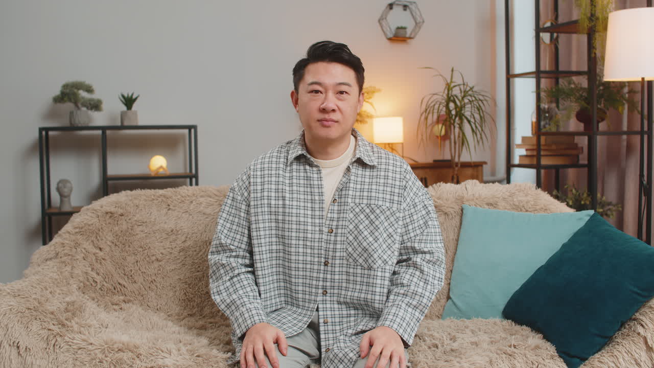 Portrait of excited smiling chinese young man showing double thumbs up sitting on sofa couch at home