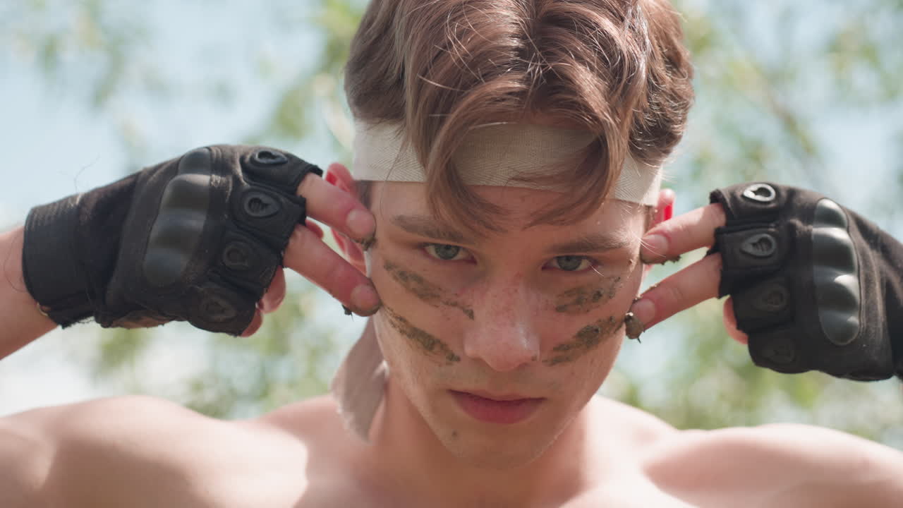 Military man wearing tactical gloves pressing wet sand between fingers, performing symbolic tiger warrior ritual in daylight, standing confidently outdoors with belt and cargo pants visible in frame