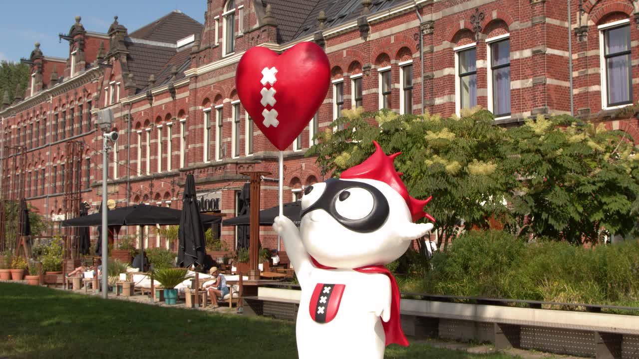 Cartoon mascot lifts heart balloon in sunny park near ornate brick building, steady camera