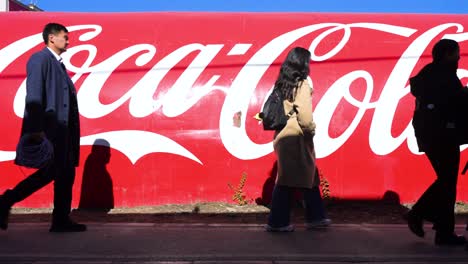 on a sunny, clear day, people pass by a wall in ulaanbaatar, mongolia, displaying the coca-cola logo and its signature red color, promoting the american soft drink brand.