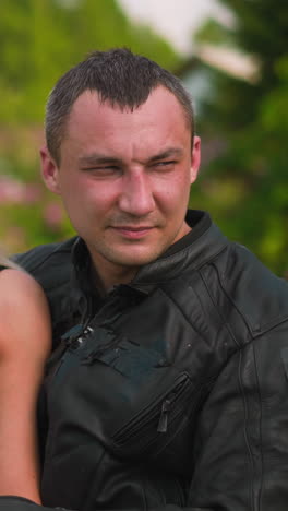 attractive woman and skilled biker man in leather moto jacket sit on motorcycle against village buildings and gardens on sunny day closeup slow motion