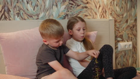 siblings sitting on bed eating fresh berries little boy plucks berry from branch drops one while showing sister playful bonding cozy bedroom scene colorful wallpaper electric socket in background