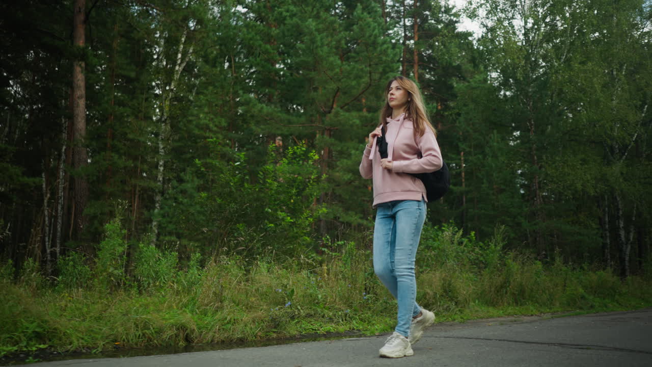 Woman in pink hoodie and jeans walking on tarred road surrounded by lush green forest, holding black shoulder bag strap confidently while passing through natural scenery on a calm day