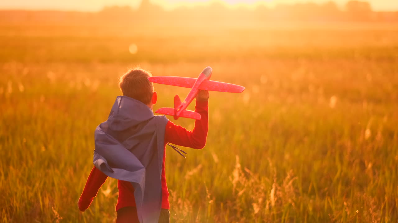 el niño con el disfraz de un superhéroe corriendo en una capa roja riendo al atardecer en el campo de verano representando que él era el piloto del avión