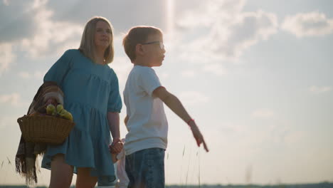 a young boy in glasses and a white shirt points excitedly towards a lake while his mother in a blue gown holds his younger sibling , under a partly cloudy sky, showcasing a moment of family bonding