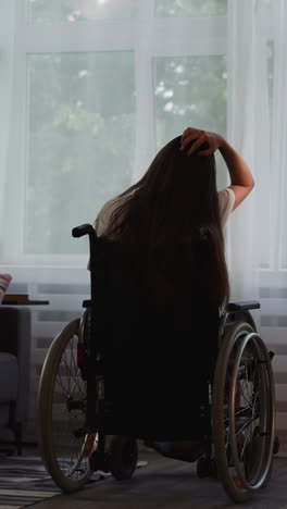 woman with disability touches long loose hair sitting in wheelchair in room. brunette female silhouette with leg injury looks out bright window backside view