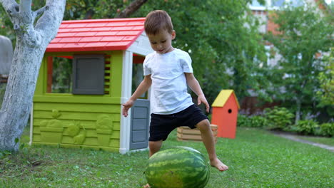 cute toddler wearing white t-shirt and black shorts sits down on the watermelon. lovely child playing outdoors in summer. playhouses at backdrop.