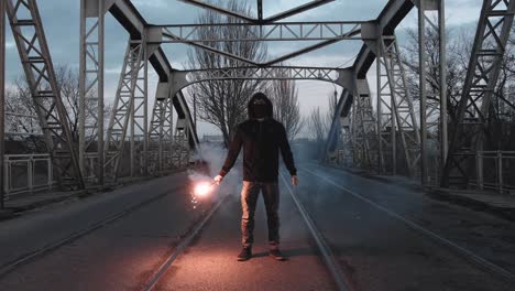 young man in hoodie and balaclava with red burning signal flare on the road under an old steel frame bridge, slow motion