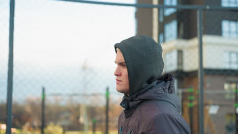 young man jogging on outdoor sport arena with focused expression, demonstrating fitness, and action, residential building backdrop and wire mesh, featuring blur view of people in background