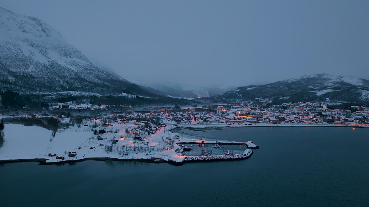 Atemberaubende Luftaufnahme eines schneebedeckten norwegischen Dorfes in der Dämmerung. Mit Hafenlichtern, nebligen Bergen und dem ruhigen Arktischen Meer auf den Lofoten.