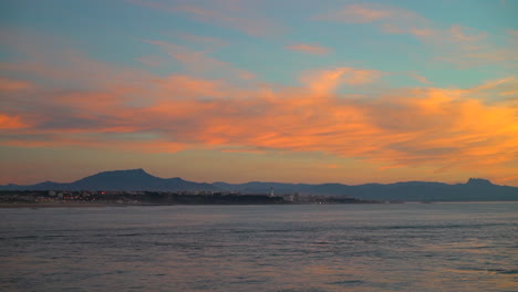 Filmisches-Eröffnungsshooting-Atemberaubende,-Immer-Noch-Dramatische-Wolken-Leuchtend-Orangefarbener-Sonnenuntergang-Abenddämmerung-Biarritz-Hossegor-Frankreich-Strand-Berg-Küstenlandschaft-Biarritz-Baskenland-Ruhiges-Wasser-Bucht-Leuchtturm-Lichter-Der-Stadt