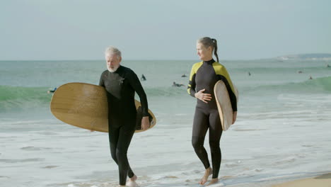 senior couple in wetsuit holding hands and walking along the beach with surfboard