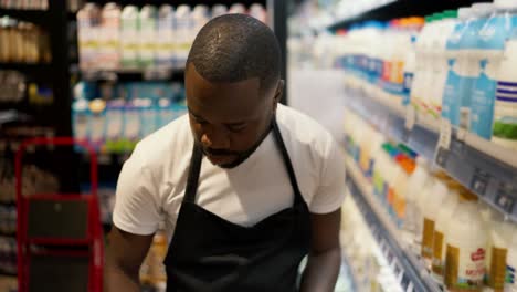 a black man in a white t-shirt and black apron laying out bottles of milk on the counter of a supermarket with dairy products