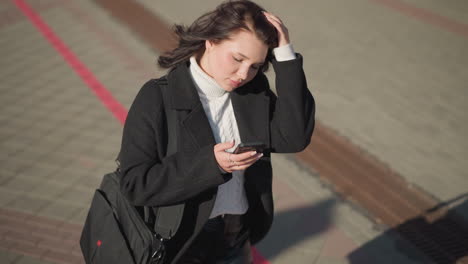 lady walking outdoors on interlocked path, touching her hair, focusing on her phone in her hand, and looking around in an urban environment while strolling in the sunlight