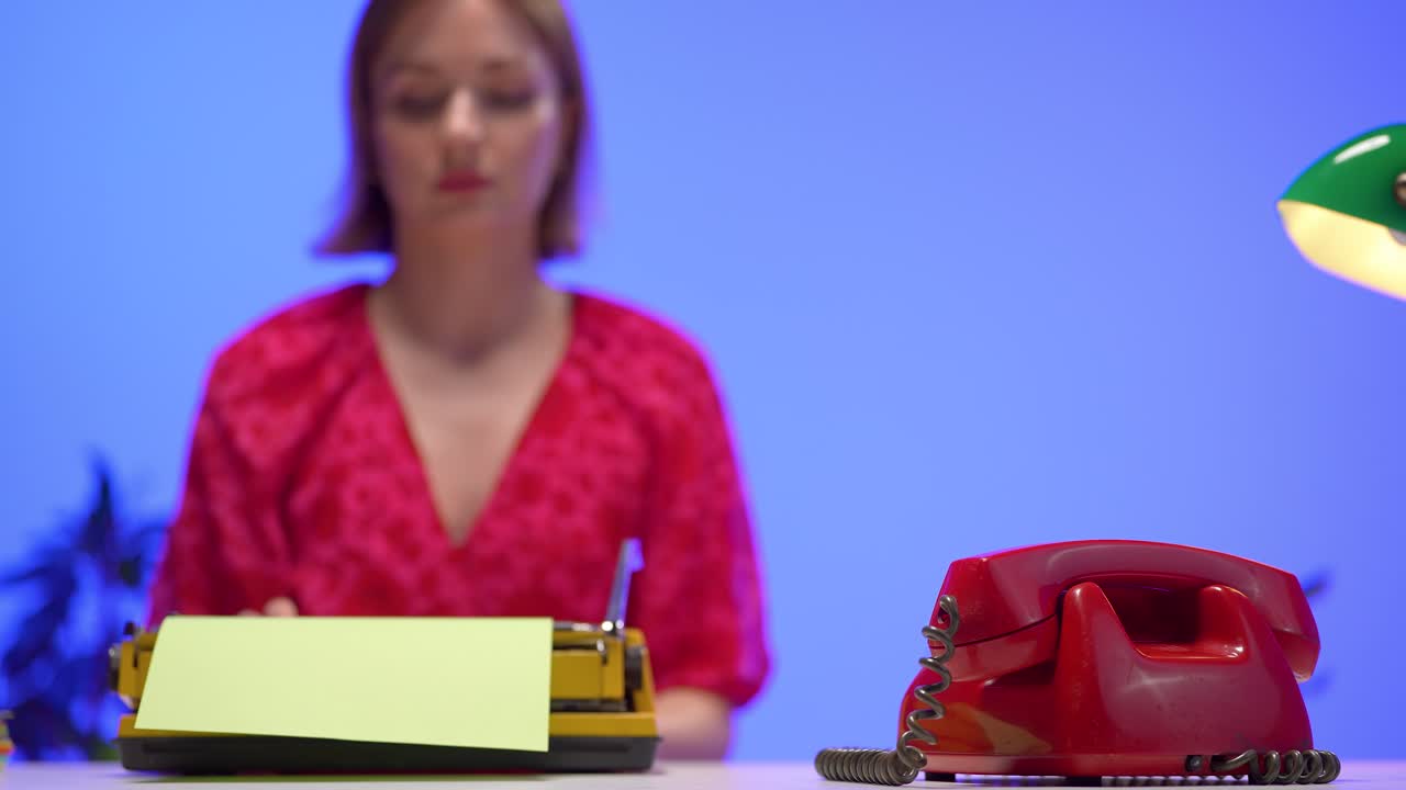 Young woman speaks into a classic red rotary phone, seated at a vintage desk with a yellow typewriter. The scene features bold retro styling against a vivid blue background and warm desk lighting