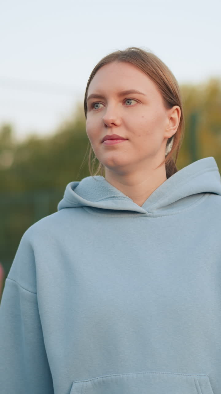 dama con capucha azul sirve voleibol con enfoque en su técnica de mano, fondo borroso de personas en entrenamiento rosa al aire libre, mostrando una sesión de práctica deportiva en una cancha de voleibol