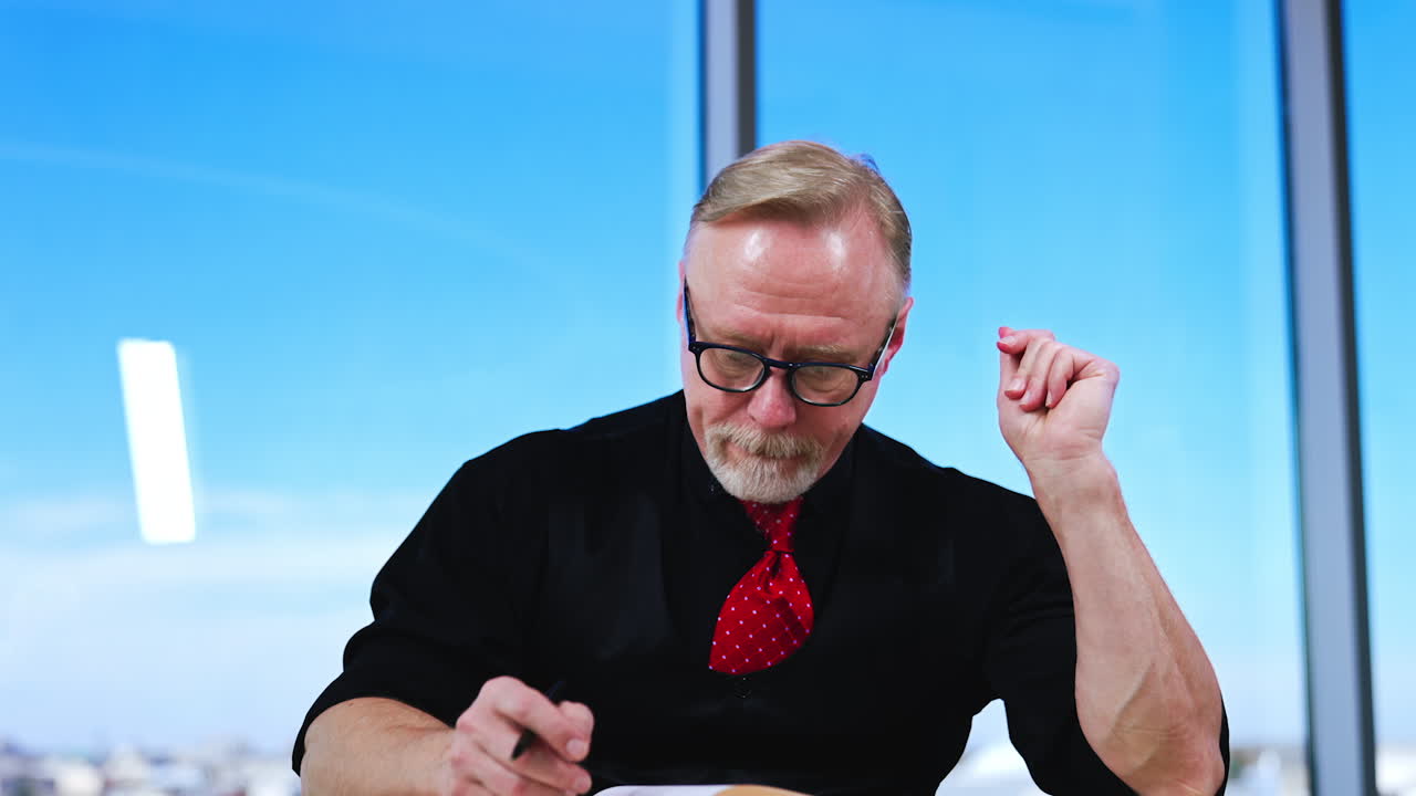 Caucasian businessman wearing glasses, black shirt, vest and red tie sits thinking over something. Man looks at paper book and then up, planning or looking for ideas.