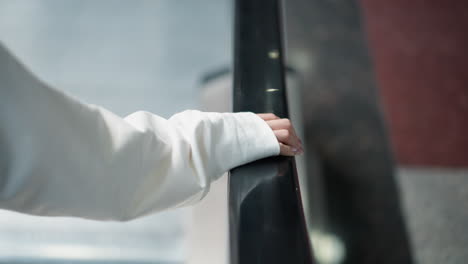 close-up of hand resting gently on shiny railing of moving escalator, showcasing polished nails, soft sleeve detail, and blurred background of modern indoor space