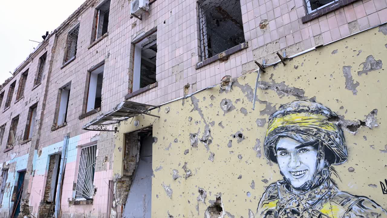 A wide shot captures a mural of a Ukrainian soldier, framed by bullet and shrapnel holes, on a heavily damaged building in Irpin, Ukraine, during the Ukraine-Russia conflict.