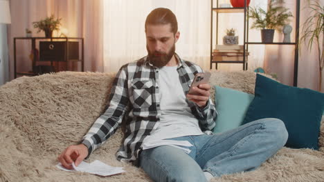 young man sorting bills at home sofa budget check phone in hand multiple papers calm focus vibe