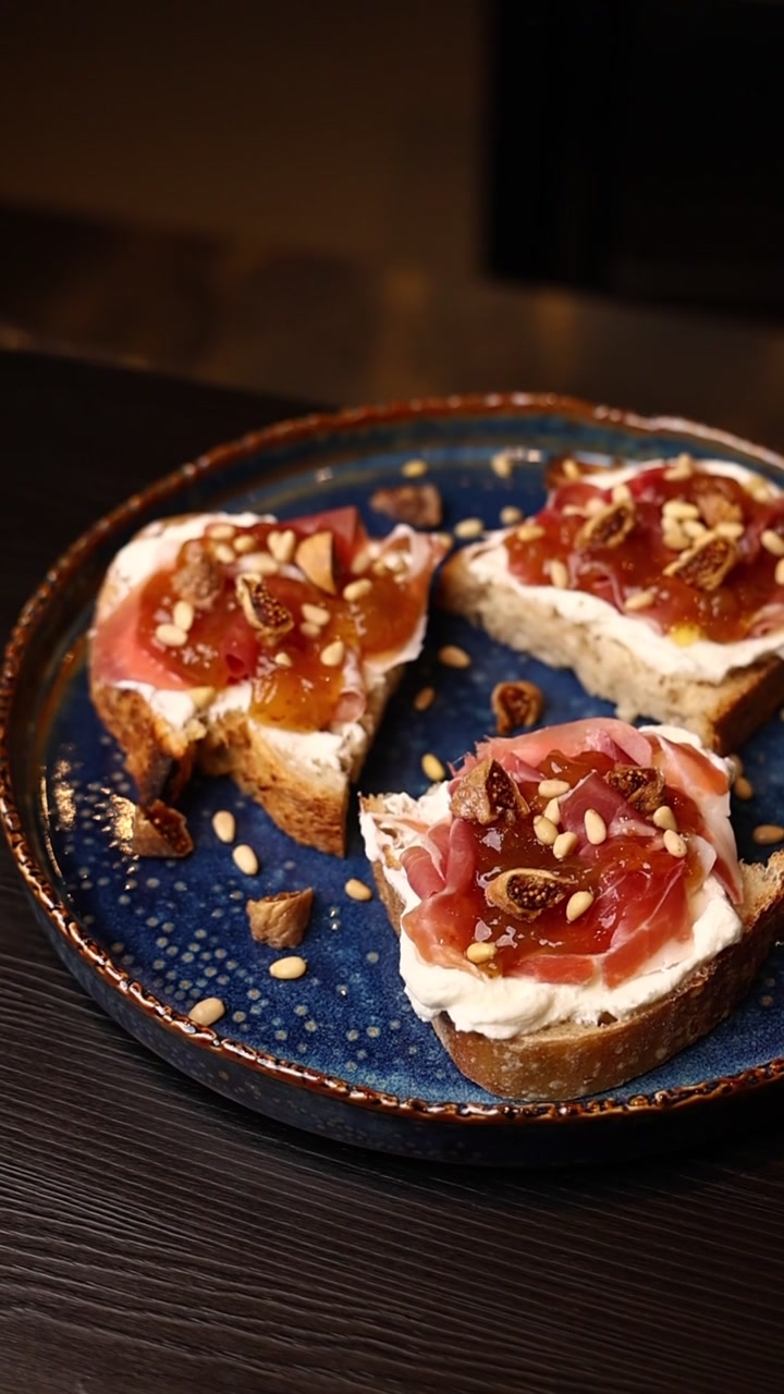 Wykwintna bruschetta z szynką parmeńską, figami i orzeszkami piniowymi na niebieskim talerzu.