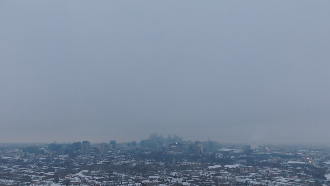 Aerial footage of a gray sky that pans down to reveal a foggy Philadelphia skyline and snow covered neighborhoods in the foreground.