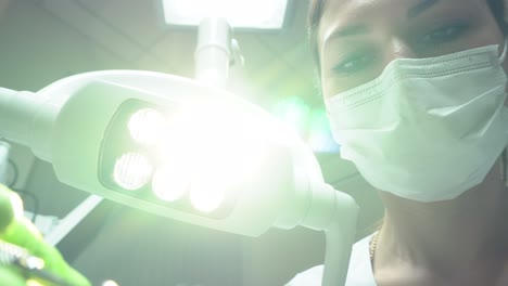 young female friendly dentist in mask approaching to the patient holding a mouth mirror and a hook with the dental lamp turned on. standing upon a patient and looking in the camera. shot in 4k