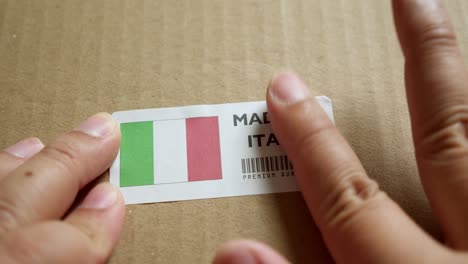 Hände-Bringen-Das-Flaggenetikett-„Made-In-Italy“-Auf-Einem-Versandkarton-Mit-Einem-Barcode-In-Premiumqualität-An