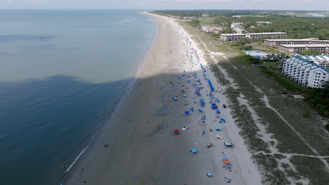 Aerial video of Hilton Head shows a long stretch of beach dotted with umbrellas, tents, and swimmers, with coastal buildings in the background