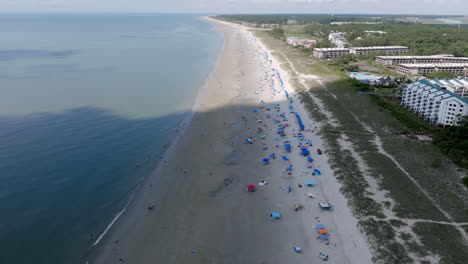aerial video of hilton head shows a long stretch of beach dotted with umbrellas, tents, and swimmers, with coastal buildings in the background