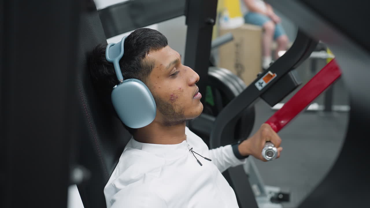 close up high school student sitting on exercise machine gripping handles while wearing wireless headset during gym workout session with other people casually seated in background