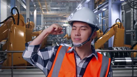 close up of asian male engineer with safety helmet flexing his bicep and smiling to camera while standing with industrial robotic arms working